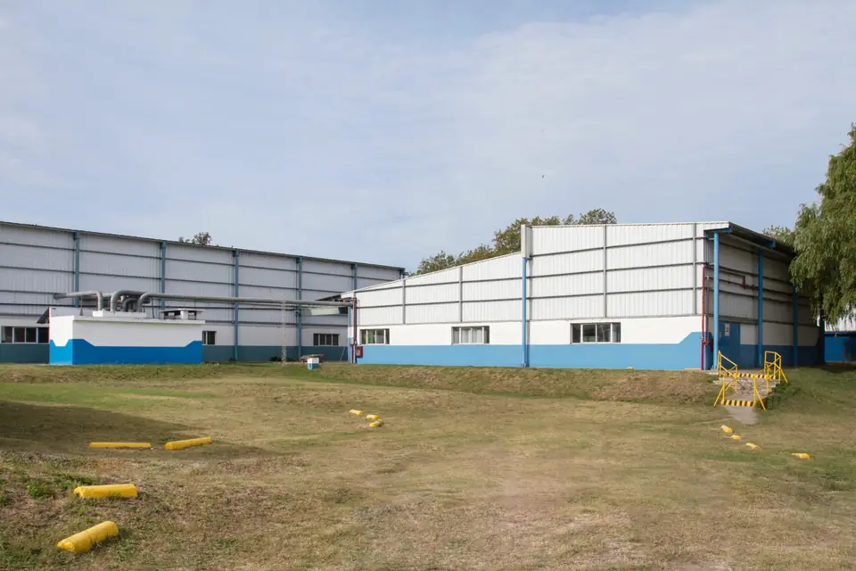Exterior of the textile manufacturing plant: two large white and blue industrial sheds with a grass yard and yellow safety markers in front.